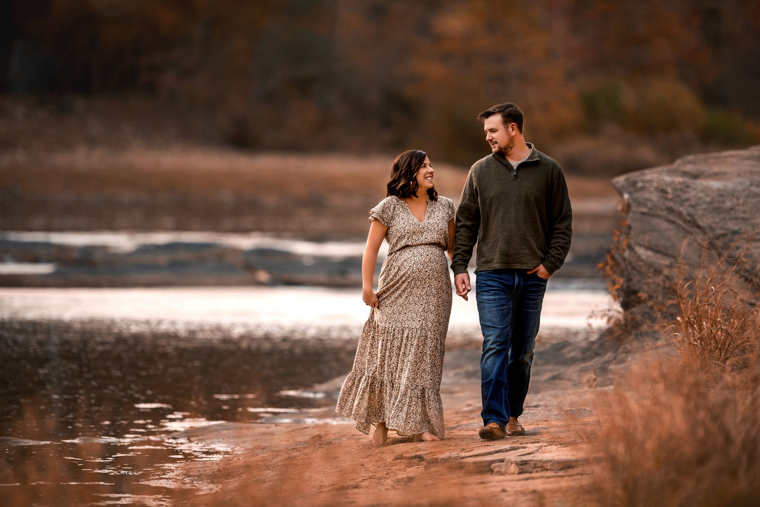 A couple walks along a scenic lakeshore during golden hour in autumn with reflections in the water.
