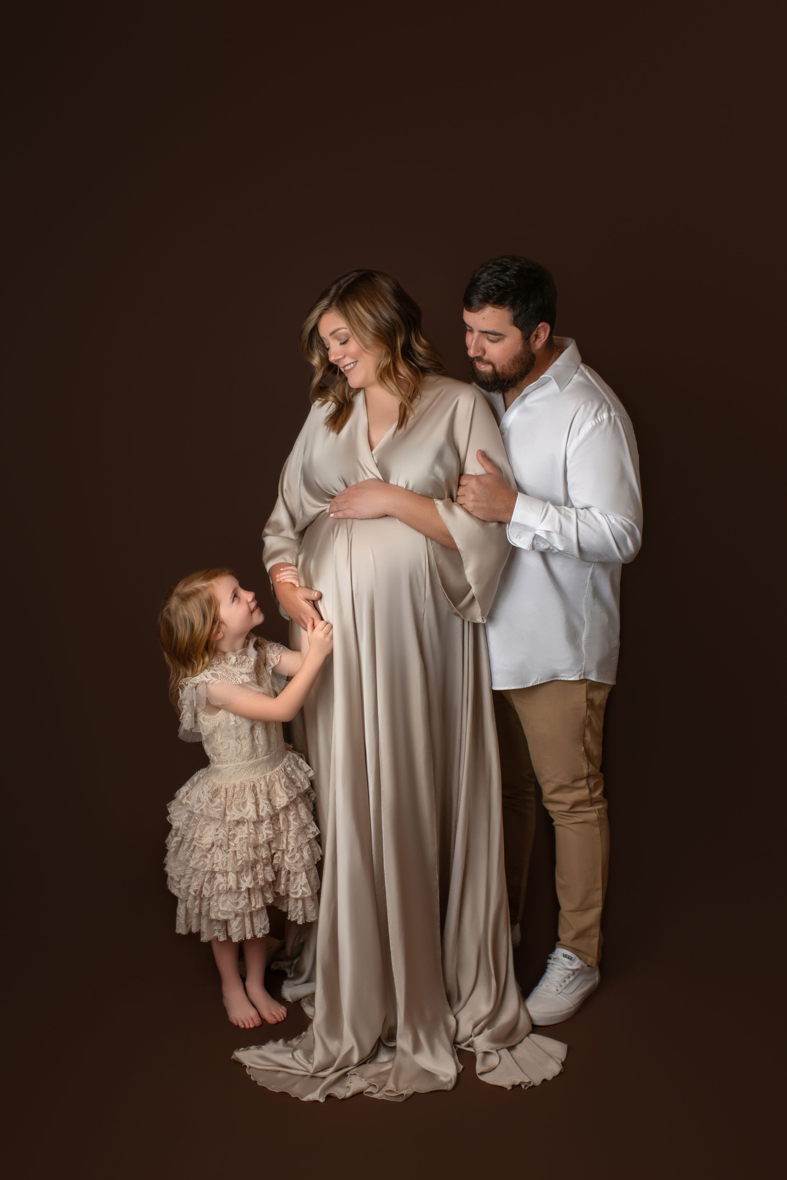 A family poses together against a dark background in elegant neutral-toned outfits for a maternity photo shoot.