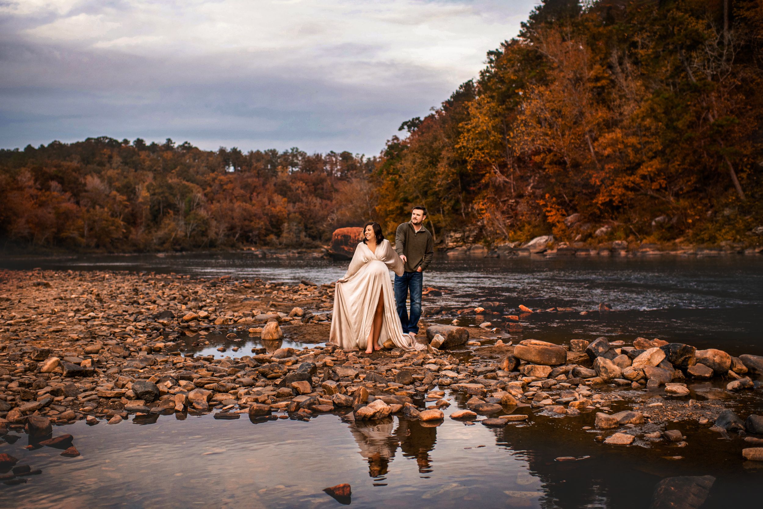 A couple stands on rocky riverbank surrounded by autumn foliage with their reflection in the water.