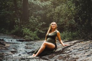 A person poses on wet rocks near a forest stream in a dark swimsuit.