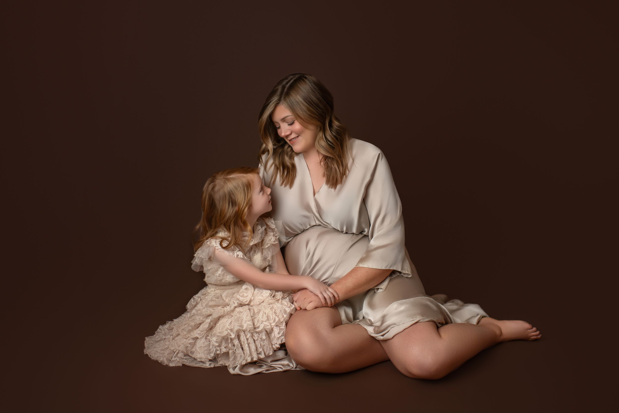 Mother and young child share a tender moment while sitting together in soft natural lighting against dark background.