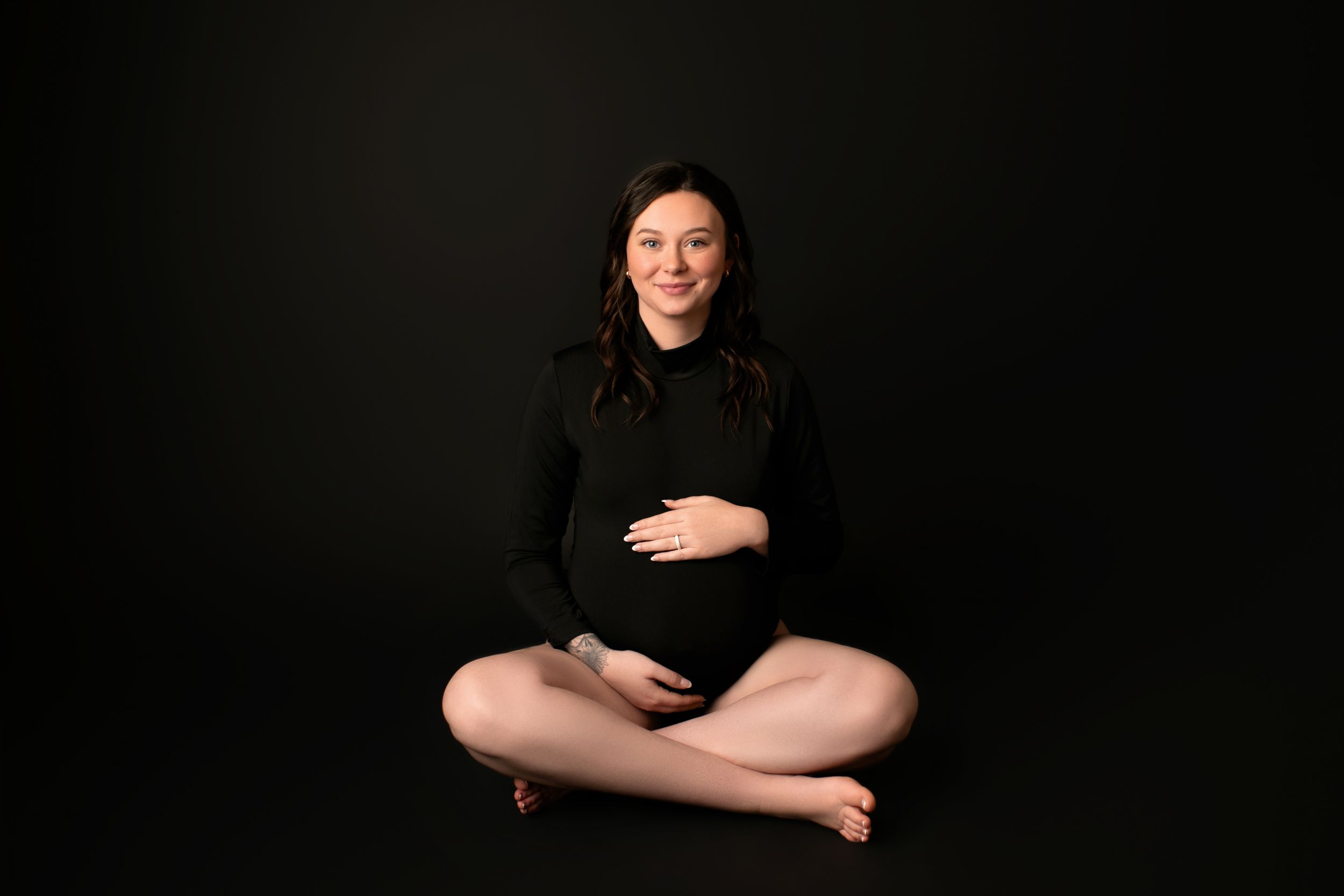 Studio maternity photo with cross-legged pose against dark background in black turtleneck sweater.