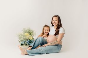 A photograph shows a mom and daughter lying on a white background in casual outfits with a gentle, candid interaction.