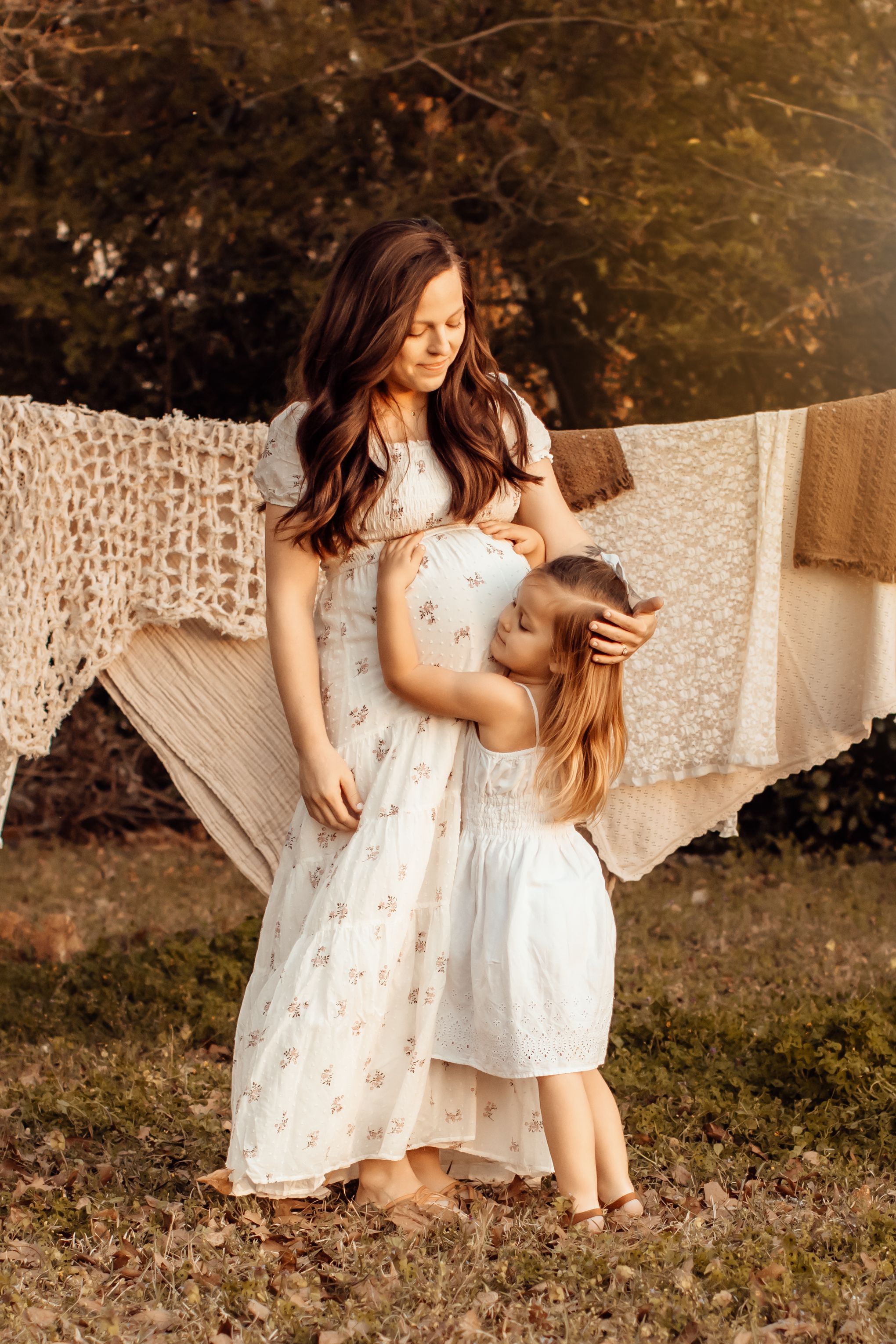 A figure in a white floral dress sits tenderly in an outdoor setting with woven lace fabric draped behind.