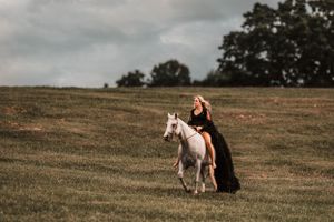 A figure in a black dress walks with a white horse across a grassy hillside.
