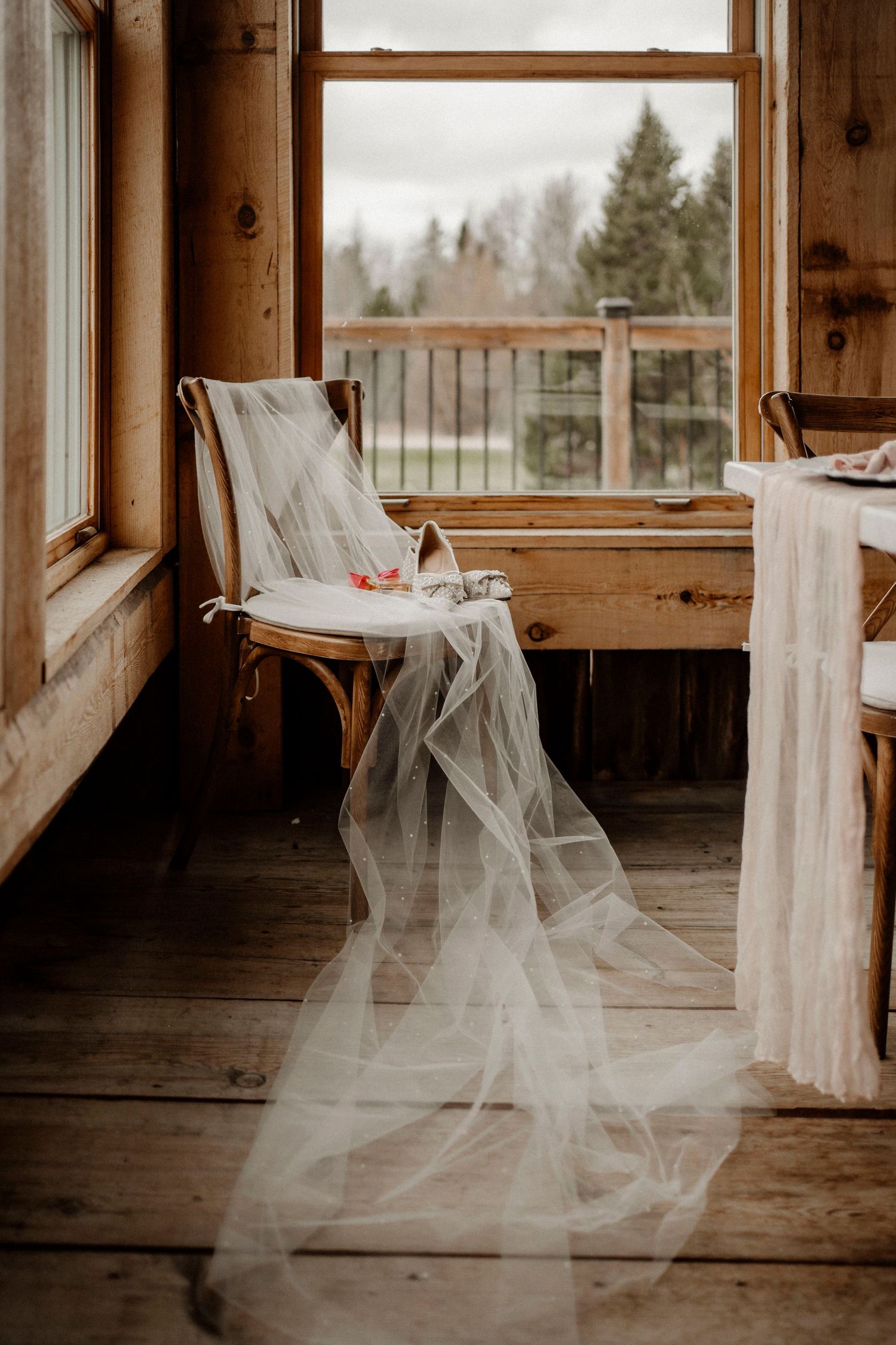 A flowing white wedding veil elegantly draped over a wooden chair by a window in a rustic cabin setting.