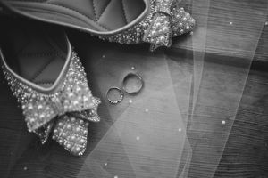 Black and white detail shot of sparkly bridal shoes with wedding rings on wooden surface.