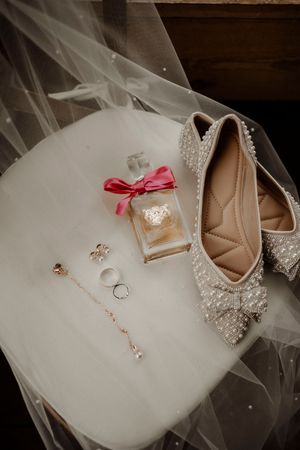 Wedding accessories including bridal shoes with crystal details, perfume with pink bow, rings and veil on white surface.