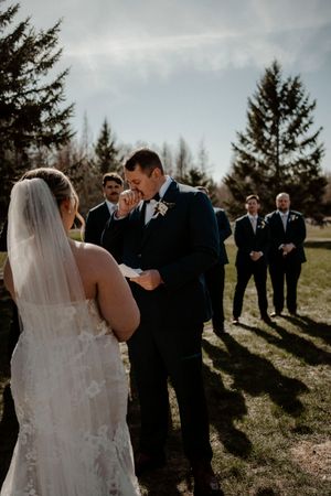 Couple exchanges vows during outdoor wedding ceremony with groomsmen standing in background on sunny day.