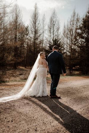 Newlyweds walk down a dirt path surrounded by tall trees during a moody autumn wedding photo session.
