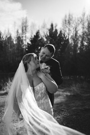 Black and white wedding portrait showing tender moment between couple outdoors with forest backdrop and flowing veil.
