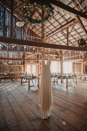 White wedding dress hanging in a rustic barn venue with wooden beams, tables, and decorative lighting.
