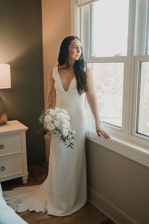Elegant bridal gown featuring a deep v-neck, sheer polka dot sleeves and a white bouquet by a window.
