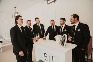A group of groomsmen in black tuxedos gather around a white bar sharing drinks and smiles at an elegant event.