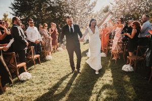 Newlyweds walk down outdoor aisle surrounded by smiling guests in sunlit garden ceremony.