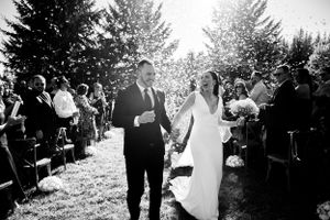 Black and white photo of a bride and groom walking down the aisle at an outdoor wedding ceremony surrounded by guests.
