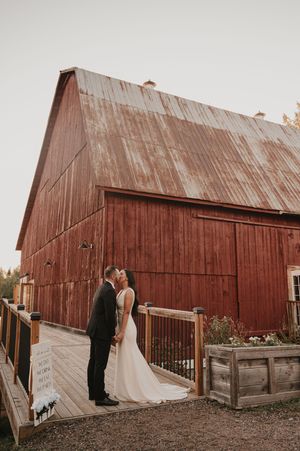 A romantic wedding moment captured in front of a rustic red barn with wooden deck and railing.