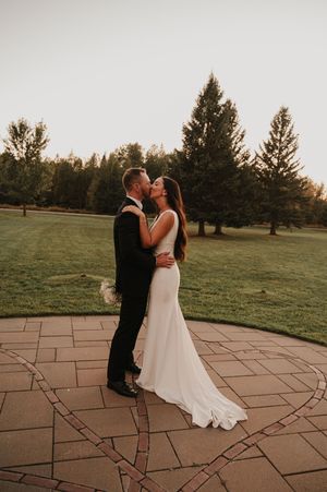 A romantic sunset wedding moment on a stone patio surrounded by evergreen trees and grassy fields.