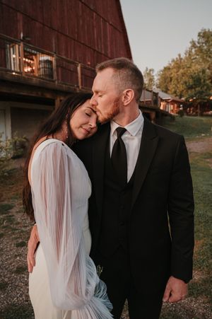 A romantic couple in wedding attire embrace by a rustic red barn during golden hour.