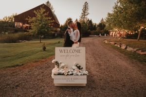 Rustic wedding welcome sign with angel wings displayed on driveway entrance near wooden barn at sunset.