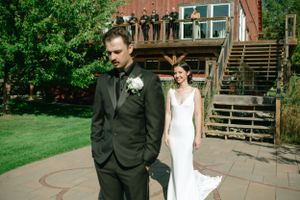 A couple in wedding attire stands on a brick patio beside wooden stairs at a rustic venue on a sunny day.