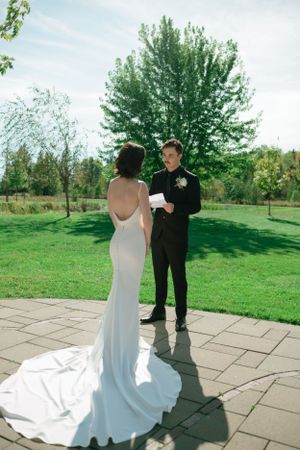 A bride in a elegant white gown and groom in black suit share a first look moment on a stone patio surrounded by greenery.