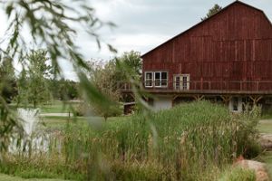 A weathered red barn sits nestled among tall grass and trees beside a small pond on a cloudy day.