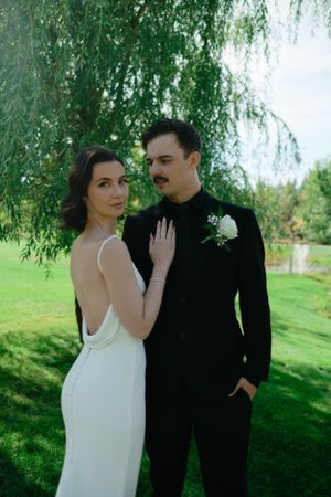 A couple poses under a weeping willow tree, the bride in a sleek white dress and groom in classic black suit.