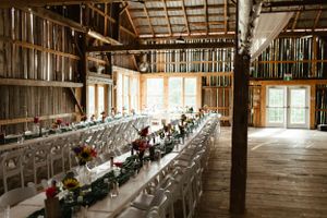 Long rustic barn wedding reception tables set with white chairs and floral centerpieces in a wooden venue with natural light.