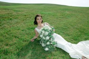 Bride in white dress poses with elegant floral bouquet on rolling green hillside.
