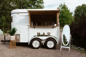 Vintage white horse trailer converted into a mobile bar with a surfboard and stool displayed outside.