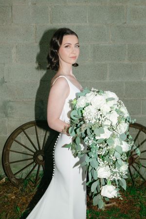 Elegant bride in fitted white dress holds cascading bouquet of white flowers and eucalyptus against stone wall.