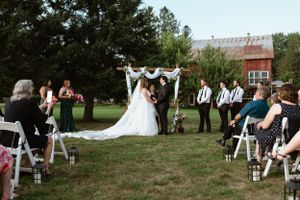 Outdoor wedding ceremony takes place on a lawn in front of a rustic red barn with guests seated on white chairs.
