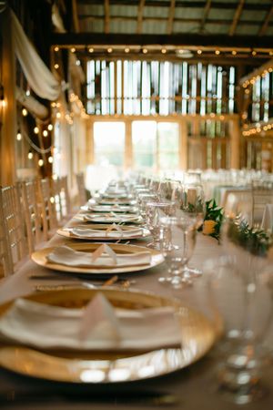 An elegant wedding reception table setting with gold chargers, white linens and twinkling lights in a rustic barn venue.