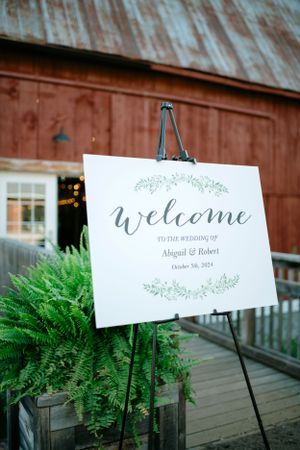 A rustic welcome sign on an easel features calligraphy text against a weathered barn backdrop with ferns.