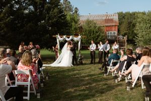 An outdoor wedding ceremony takes place in a garden setting with white folding chairs and a rustic barn in the background.