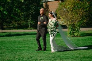 A couple in formal wedding attire walk together across a lush green lawn with shrubs and trees in the background.