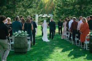 An outdoor wedding ceremony takes place on a beautiful lawn surrounded by evergreen trees and seated guests.