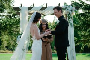 An outdoor wedding ceremony takes place under a white draped arch with greenery in the background.