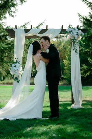Wedding ceremony under wooden arch with white fabric and floral decorations in outdoor garden setting.