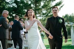 Wedding party exits ceremony on outdoor path surrounded by evergreen trees.