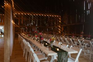 Long banquet tables with white chairs set up in a rustic barn venue with string lights hanging from wooden beams overhead.