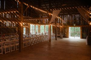 Rustic barn interior decorated with string lights and white chairs set up for a wedding reception or event.