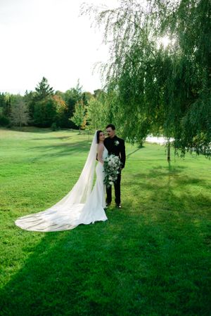 A wedding couple stands beneath a drooping willow tree on lush green grass during an outdoor summer ceremony.
