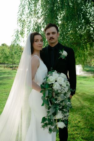 A wedding couple poses under a weeping willow tree with a romantic white floral bouquet in garden setting.
