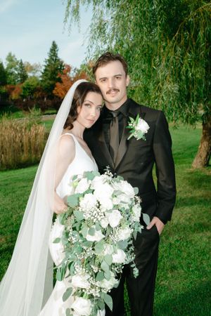 A wedding couple poses under a weeping willow tree with a cascading white floral bouquet and elegant attire.