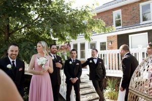 Wedding party members gather on a brick house porch with white railing on a sunny afternoon.