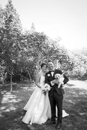 A black and white wedding photo in a garden setting featuring a couple holding their small white dog.