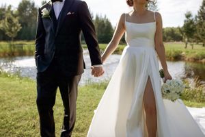 A couple in wedding attire holding hands while walking outdoors near a pond on a sunny day.