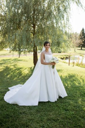 A bride in an elegant white ballgown wedding dress stands beneath a weeping willow tree on a sunny summer day.